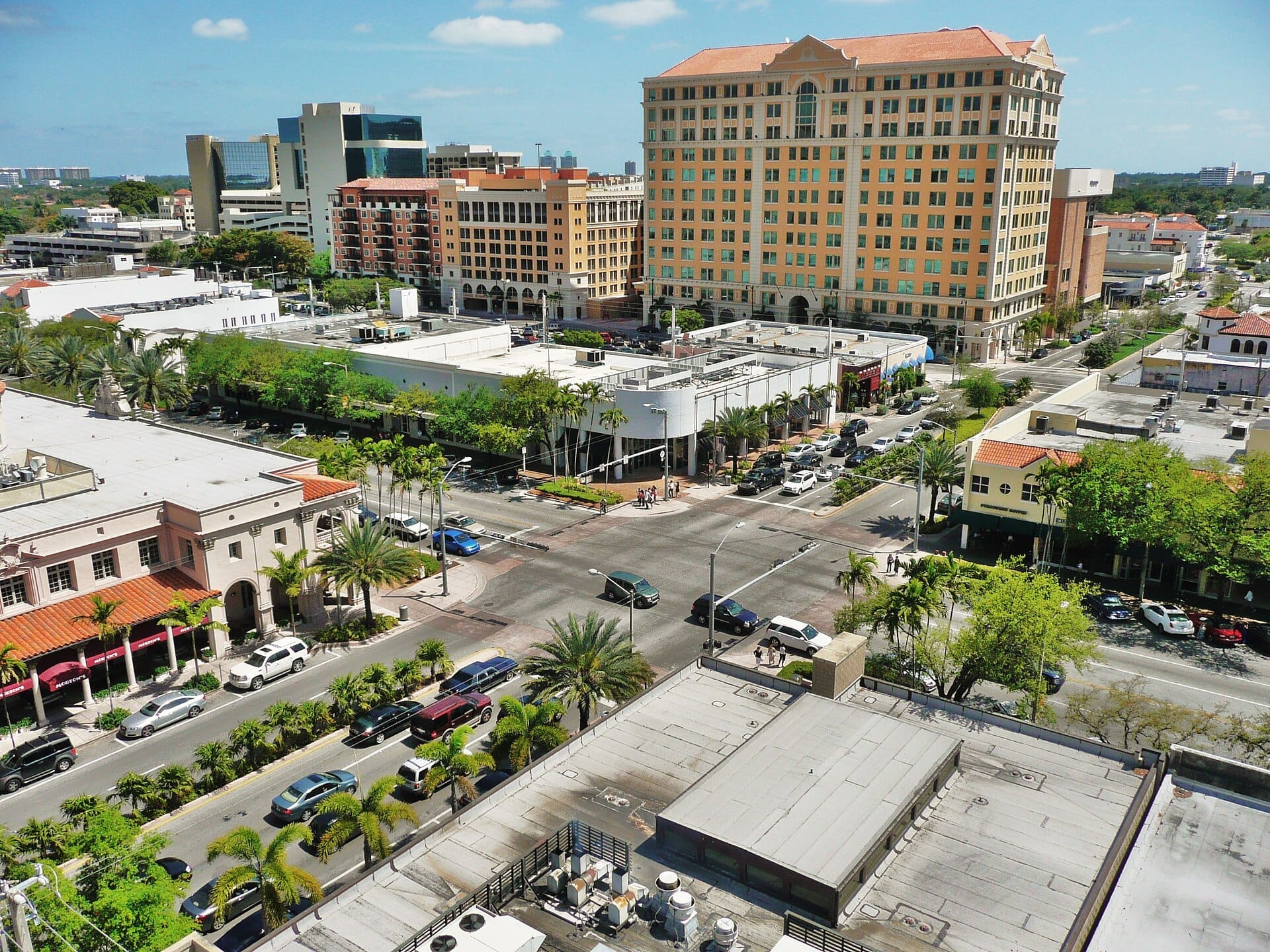 Vista aerea di Miracle Mile a Coral Gables con palme, edifici in stile mediterraneo, strade trafficate e un prominente grattacielo color sabbia sotto cieli soleggiati.