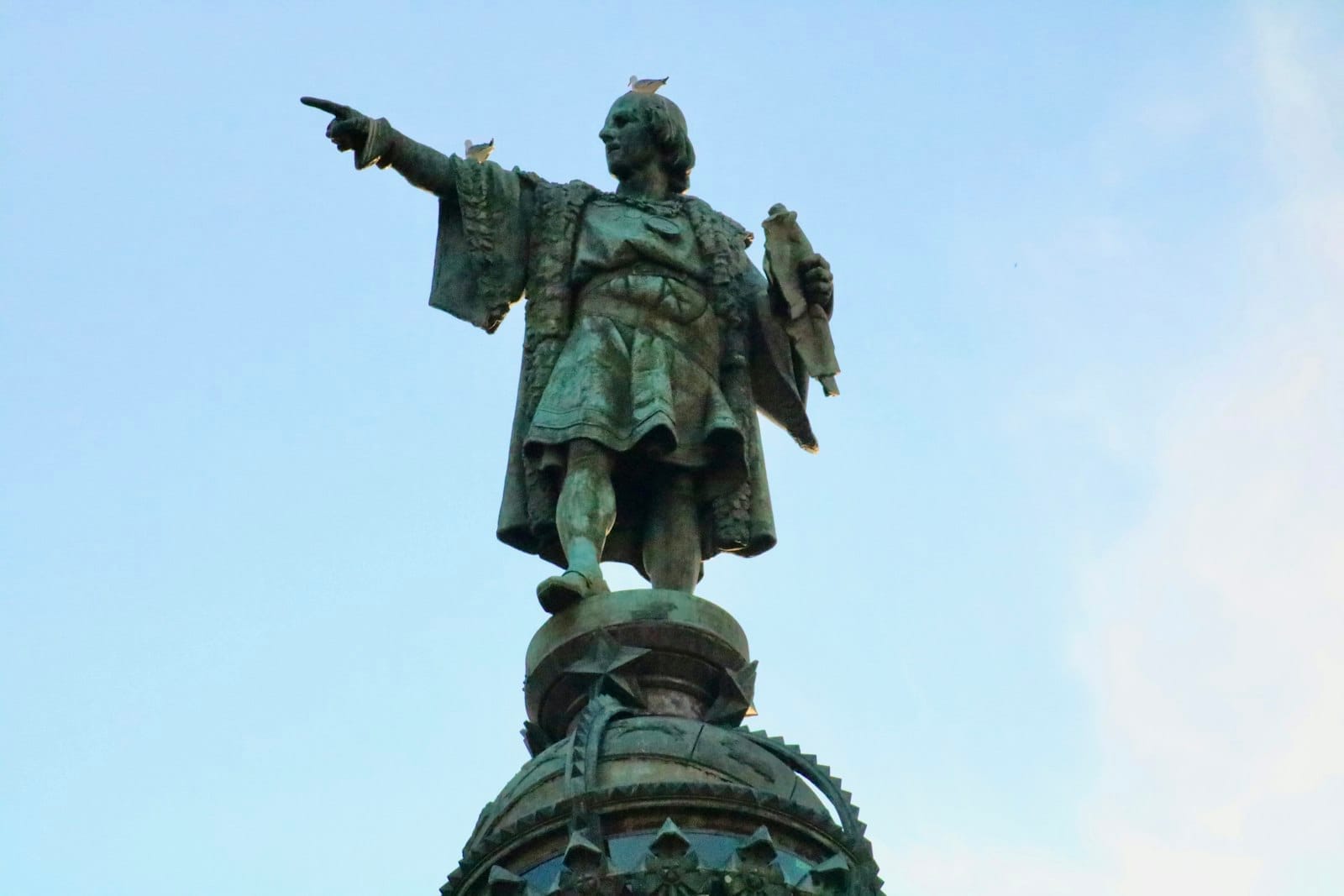 Close-up of the Columbus Monument at Mirador de Colom, bronze statue pointing skyward against a clear blue Barcelona sky.