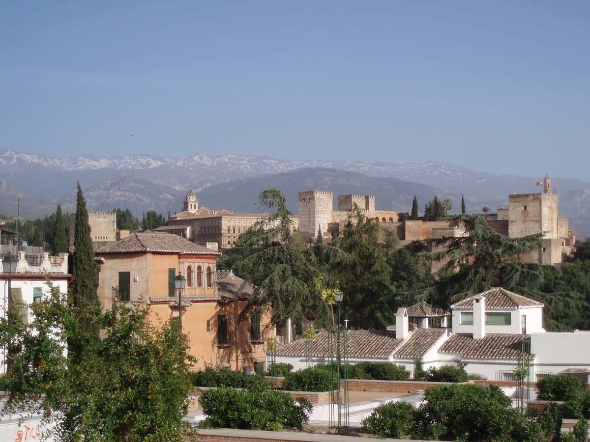 วิวจาก Mirador de San Cristóbal ที่แสดงให้เห็นหลังคาเมือง Granada ต้นไม้เขียวขจี ป้อมปราการ Alhambra และยอดเขา Sierra Nevada ที่ปกคลุมด้วยหิมะในระยะไกลใต้ท้องฟ้าแจ่มใส