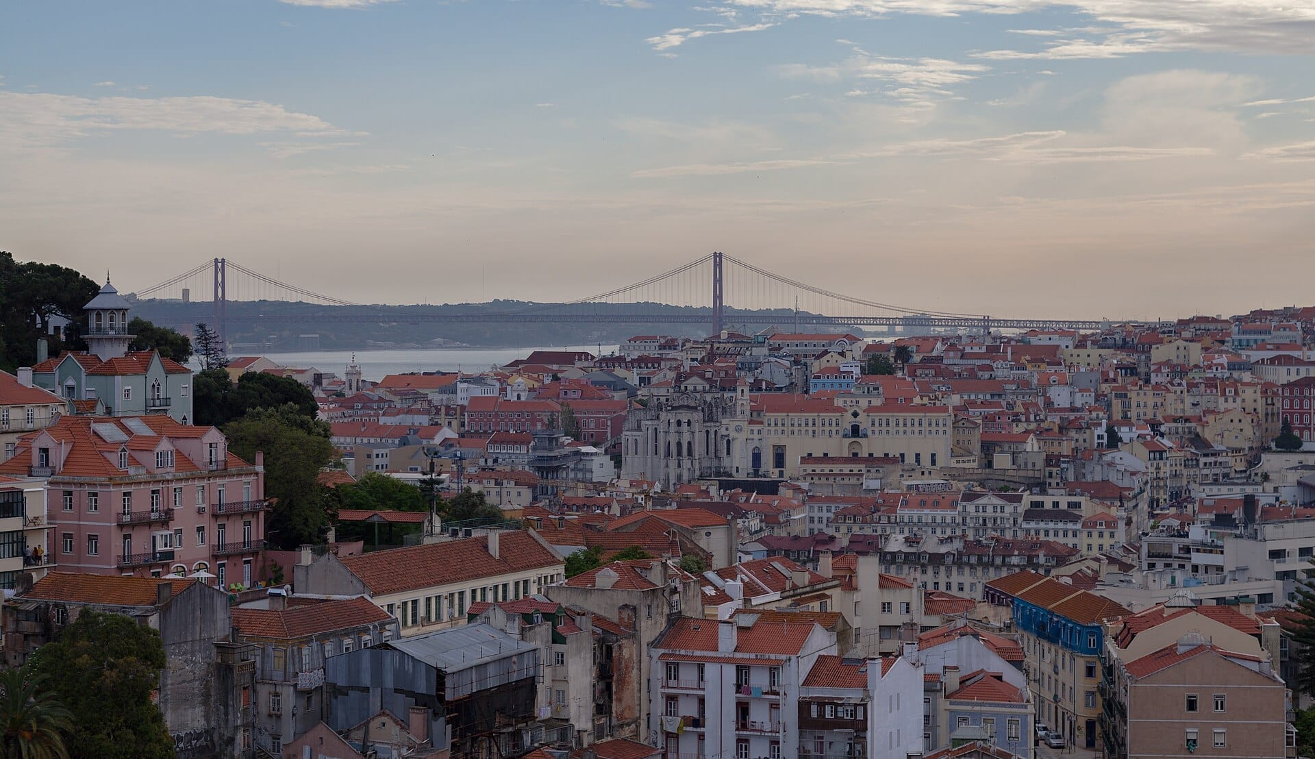 Il Miradouro da Graça con vista sui tetti rossi di Lisbona, gli edifici storici, il fiume Tago e l'iconico Ponte 25 de Abril al tramonto.