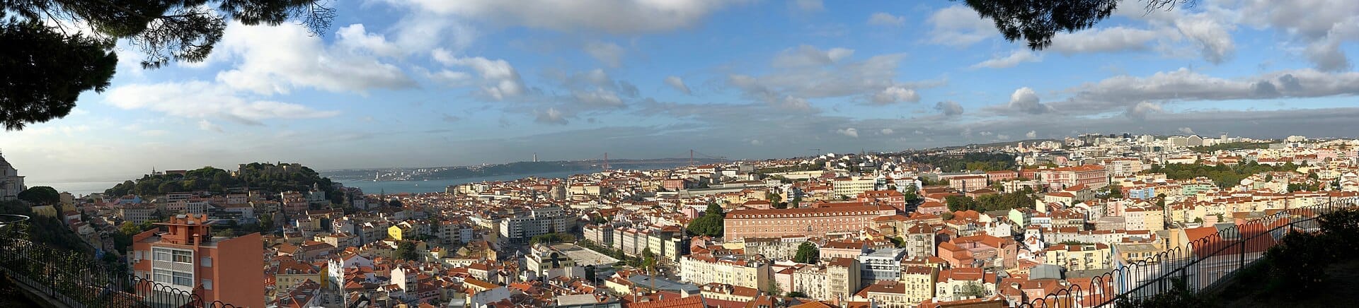 Ampia vista panoramica dal Miradouro da Senhora do Monte con i tetti di Lisbona, il Castelo de São Jorge, il fiume Tago e i ponti in lontananza sotto un cielo parzialmente nuvoloso.