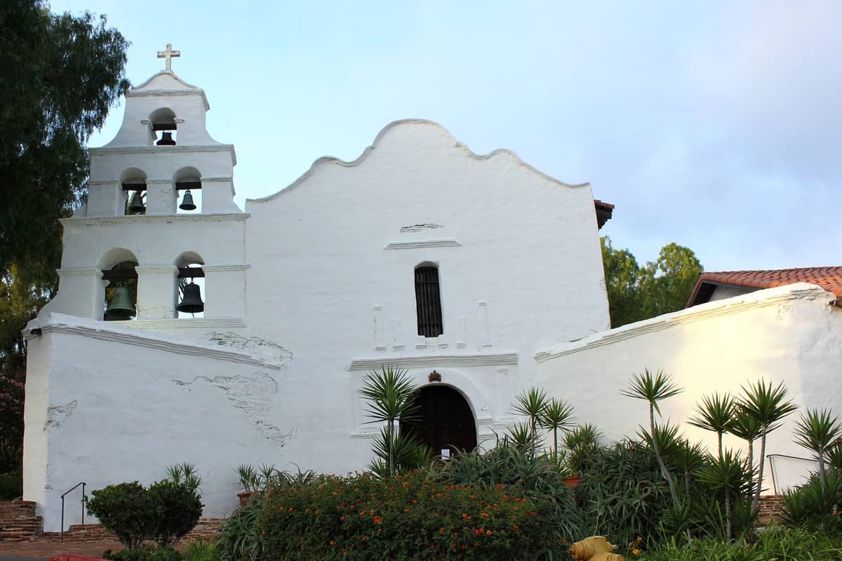 ภาพโบสถ์สีขาวกับหอระฆังอันโดดเด่นของ Mission Basilica San Diego de Alcalá รายล้อมด้วยพรรณไม้ในสวนใต้ฟ้าแจ่มใส