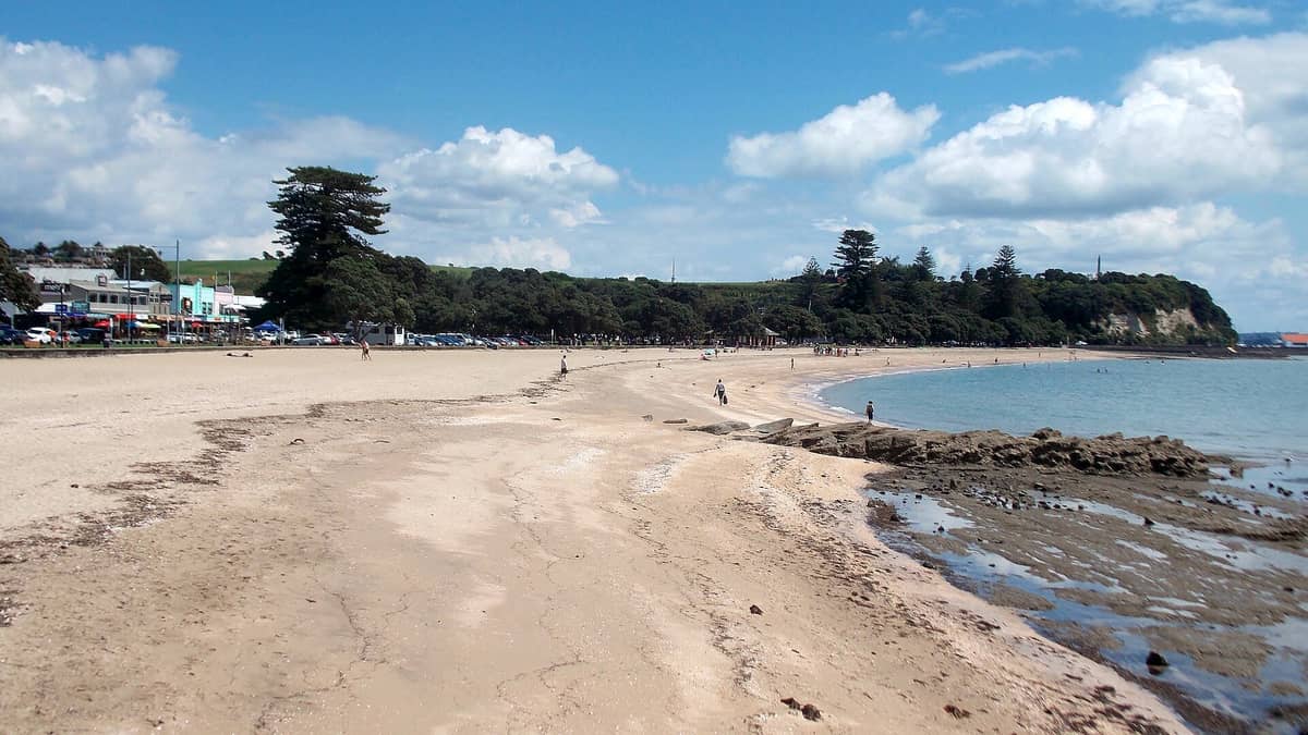 Weitläufiger Blick auf den Mission Bay Beach in Auckland mit Sandstrand, Promenade, Spaziergängern, Geschäften, geparkten Autos, Bäumen und einem Vorgebirge in der Ferne unter blauem Himmel.