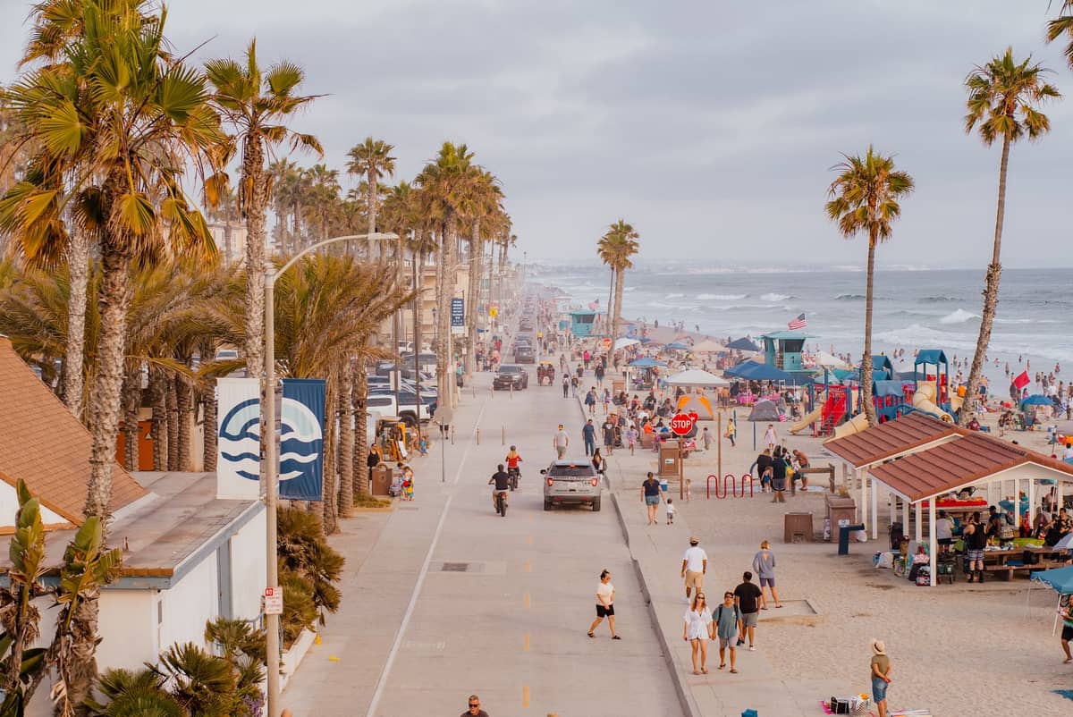 วิวชีวิตชีวา แดดสดใสของ Mission Beach Boardwalk ในซานดิเอโก คนแน่นขนัด ต้นปาล์ม ร้านค้าริมชายหาด เดินเล่น ปั่นจักรยาน พักผ่อนริมทะเล
