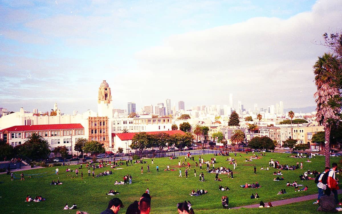 Vista del Dolores Park en el Mission District con grupos de personas descansando en el césped, edificios históricos y el horizonte de San Francisco al fondo.