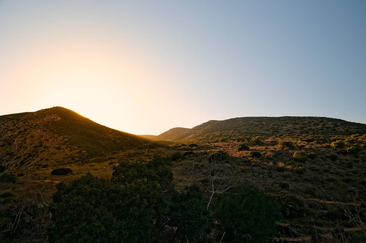 Sonnenuntergang hinter chaparral-bedeckten Hügeln im Mission Trails Regional Park – warmes goldenes Licht über der rauen Landschaft unter strahlend blauem Himmel.