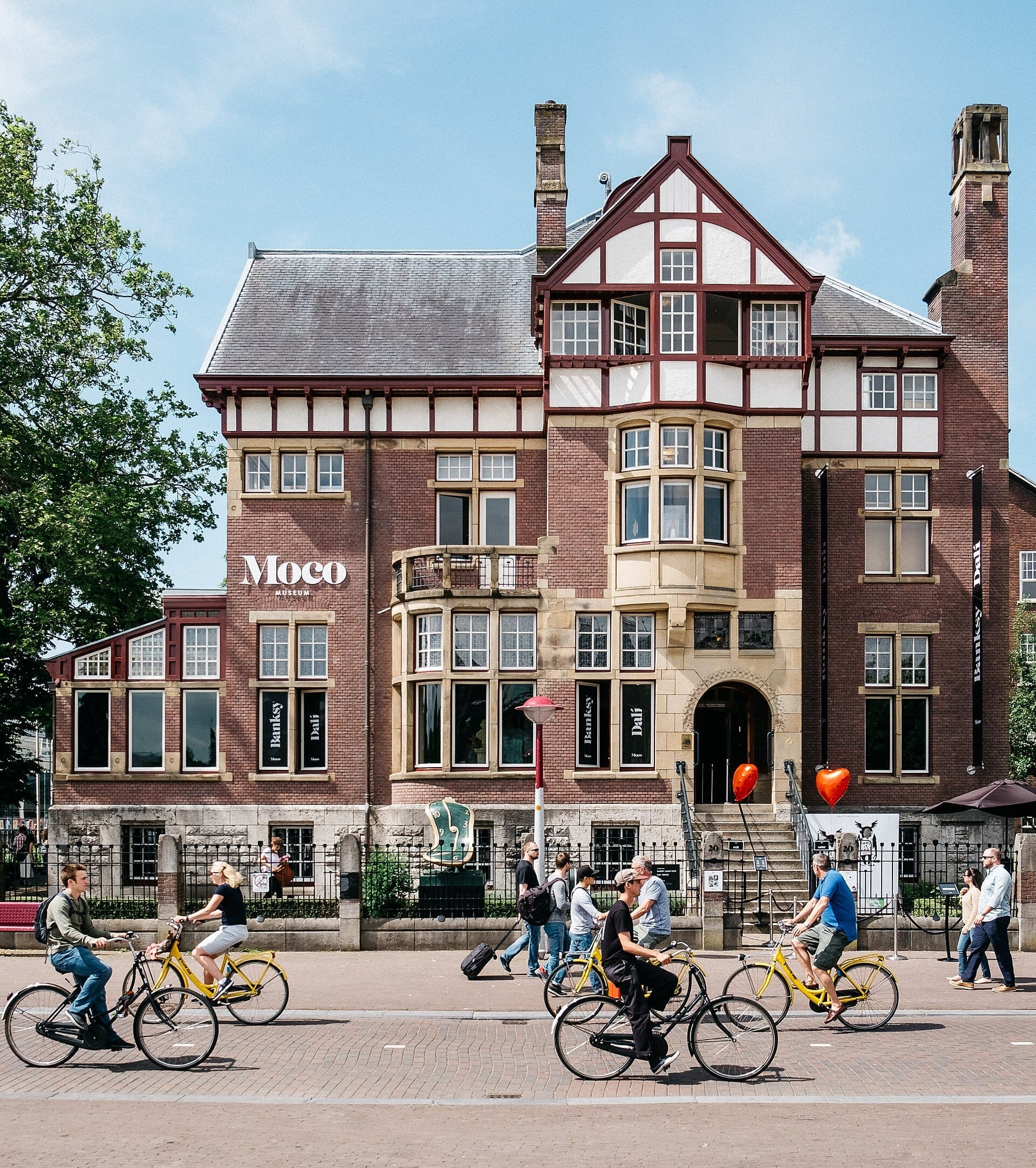 Moco Museum Amsterdam, một biệt thự gạch lịch sử với những cửa sổ lớn, hiện ra phía sau những người đi xe đạp và người đi bộ trong một ngày nắng đẹp gần Museumplein.