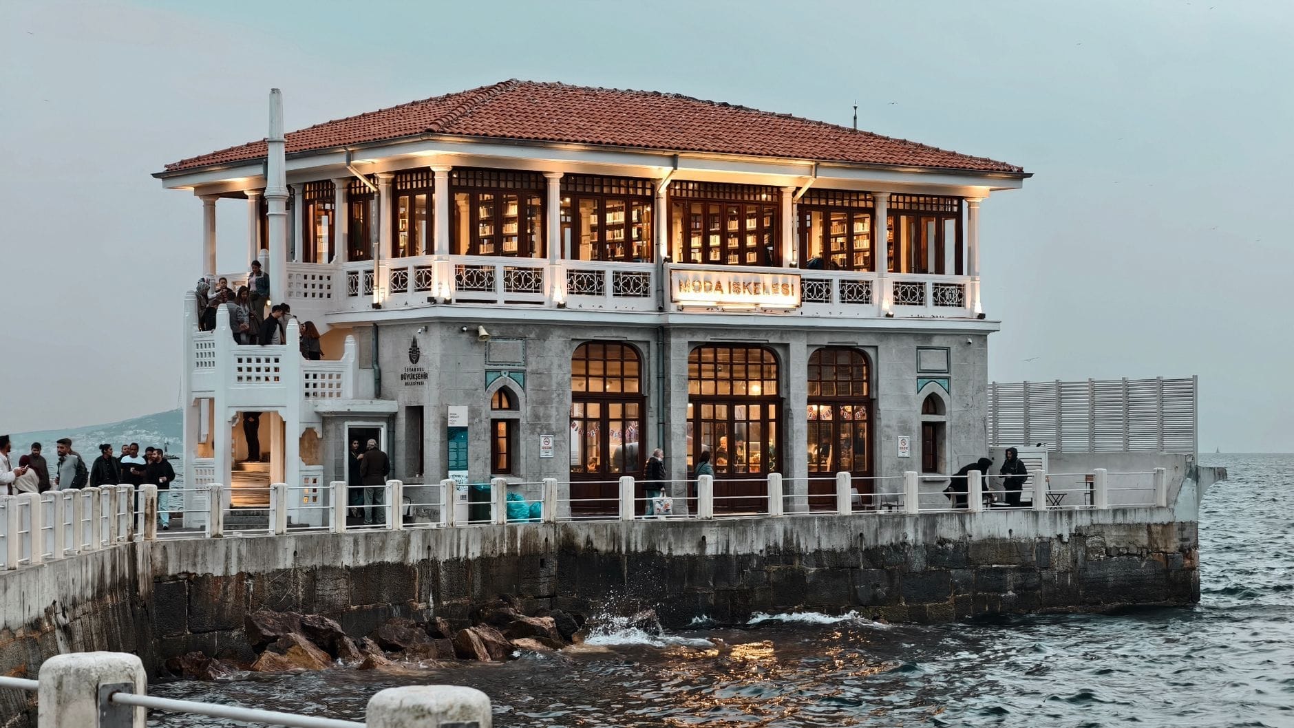 Il restaurato Molo storico di Moda illuminato calorosamente al tramonto, con persone che socializzano sul lungomare, onde che si infrangono sugli scogli e un cielo serale soffuso sul Mar di Marmara.