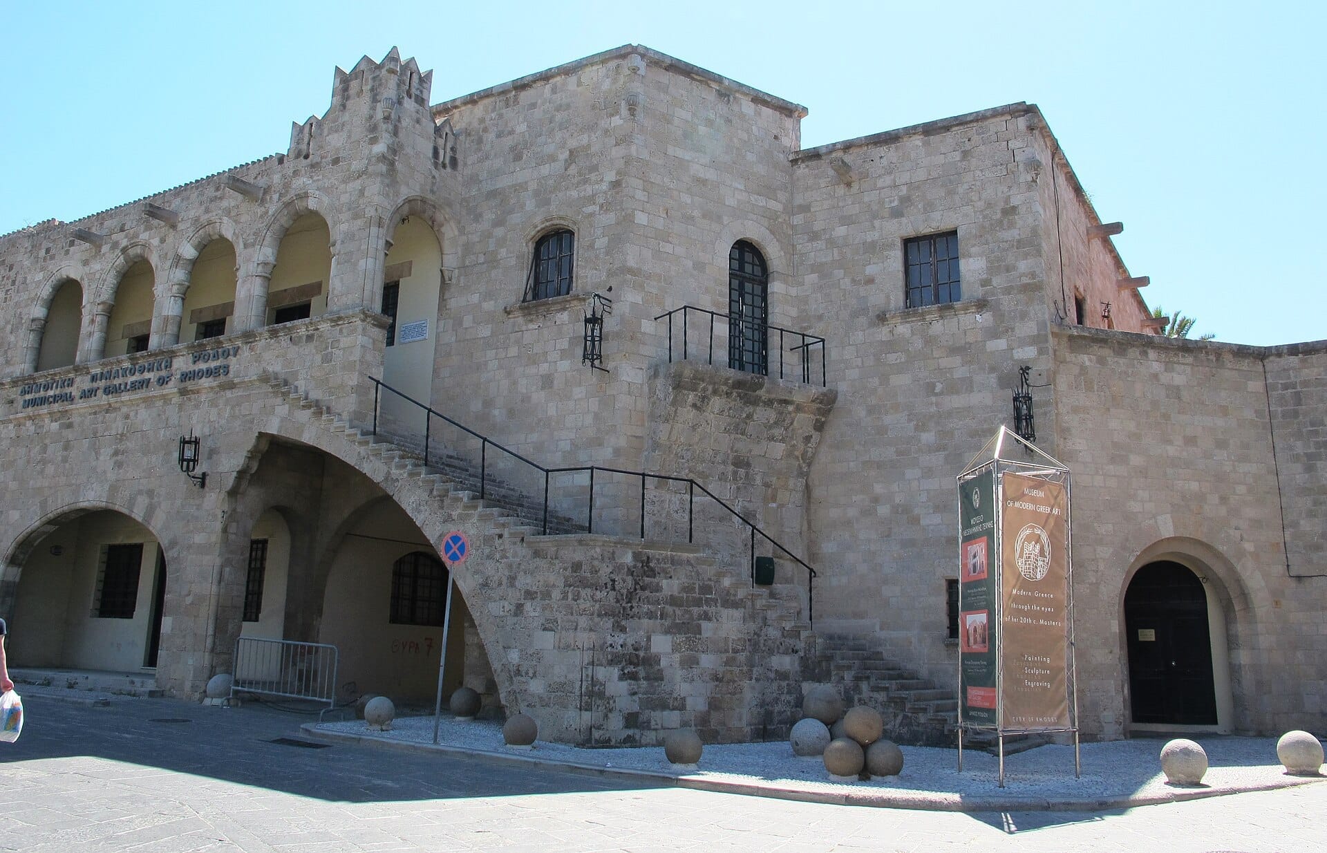 ด้านหน้าหินของ Museum of Modern Greek Art ใน Rhodes มีหน้าต่างโค้ง บันไดภายนอก และป้ายพิพิธภัณฑ์ภายใต้แสงแดดสดใส