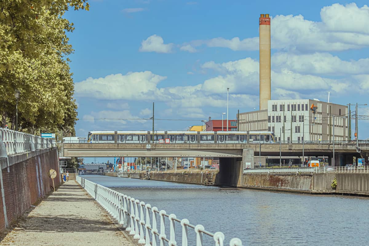Vista do Canal de Bruxelas em Molenbeek com um trem de metrô cruzando uma ponte, galpões industriais e árvores às margens do canal sob um céu azul.