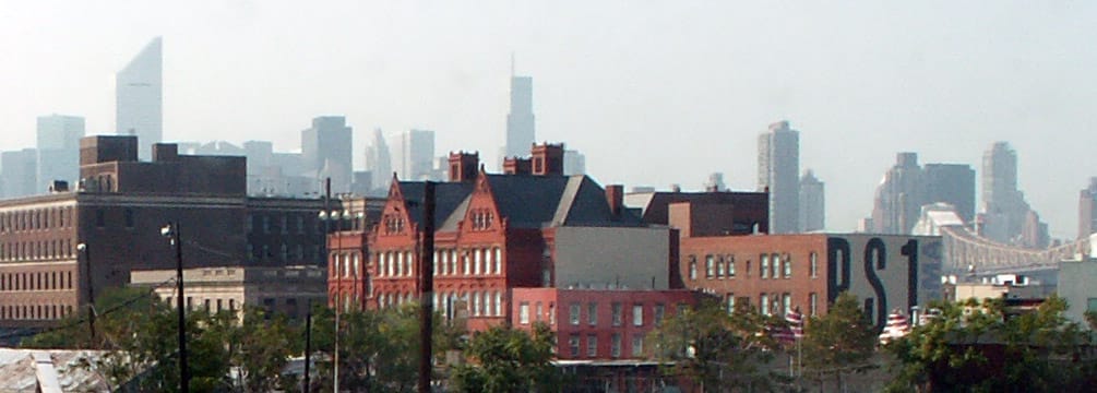 Lo storico edificio in mattoni rossi del MoMA PS1 si erge tra le strutture industriali del Queens con lo skyline di New York City visibile sullo sfondo.