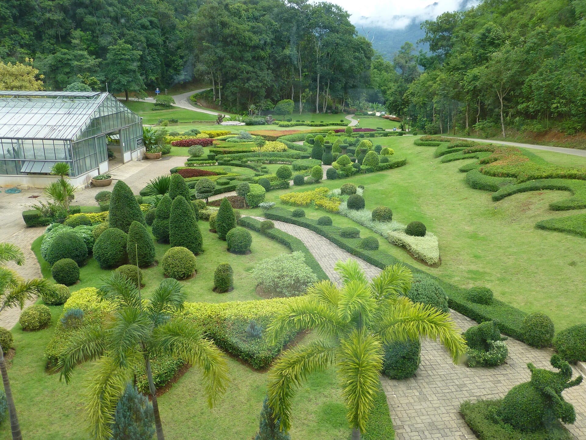 Những khu vườn được chăm sóc kỹ lưỡng và nhà kính bằng kính tại Mon Cham, Chiang Mai, bao quanh bởi những ngọn đồi xanh tươi và những con đường đá uốn lượn dưới bầu trời nhiều mây.