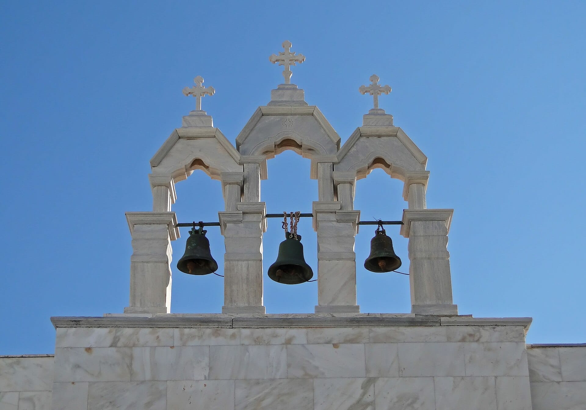 Torre campanario de mármol blanco con tres campanas y cruces en el Monasterio de Panagia Tourliani, con el despejado cielo azul de Mykonos de fondo.