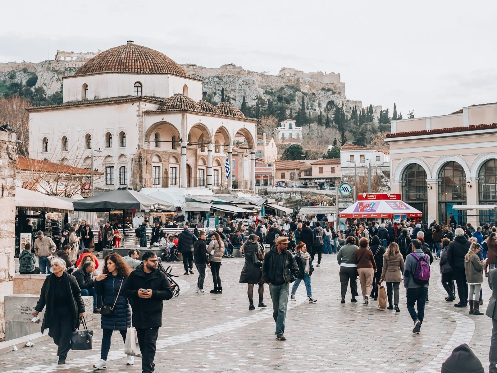 Folle esplorano il Mercato delle Pulci di Monastiraki ad Atene, circondate da edifici storici e bancarelle, con l'Acropoli visibile sullo sfondo.