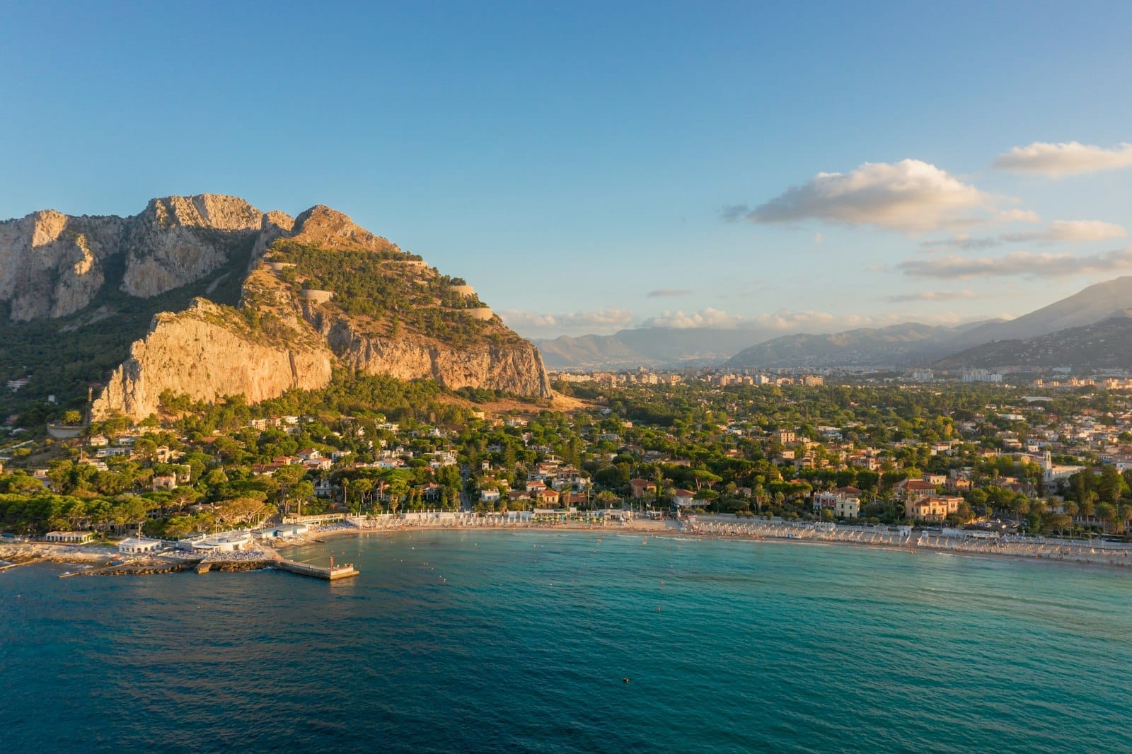 Luftaufnahme des Strandes von Mondello mit hellem Sand, türkisfarbenem Wasser und üppigem Grün, eingerahmt vom Monte Pellegrino bei Sonnenuntergang nahe Palermo.