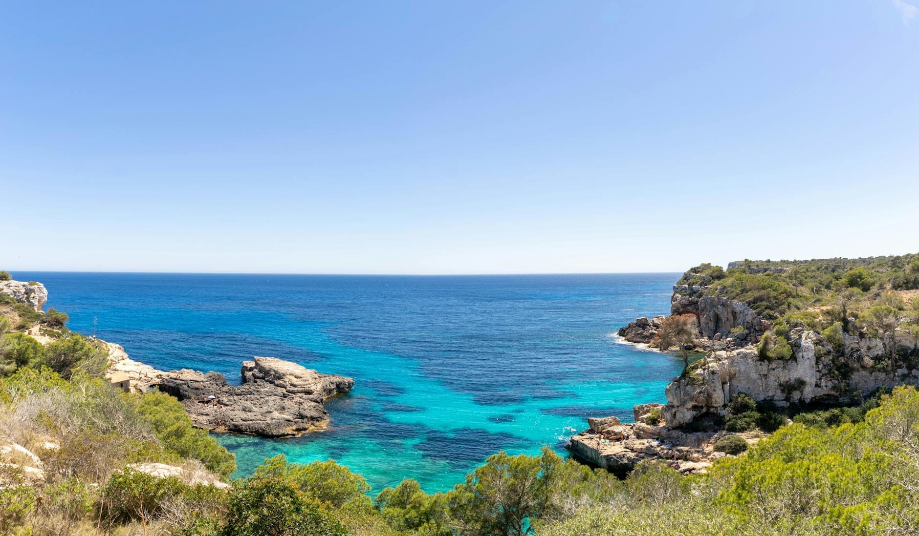 Acque cristalline turchesi incorniciate da scogliere rocciose e una fitta vegetazione verde sotto un cielo azzurro e soleggiato, sulla costa protetta del Parco Naturale di Mondragó.