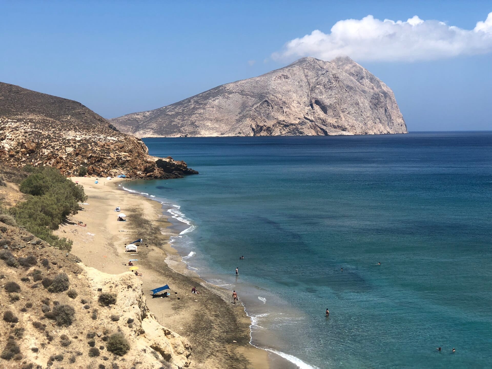 หาด Monolithos ที่มีชายฝั่งทรายสีดำ นักอาบแดดไม่กี่คน คลื่นเบาๆ น้ำสีฟ้า หน้าผาหิน และภูเขาขนาดใหญ่เป็นฉากหลัง