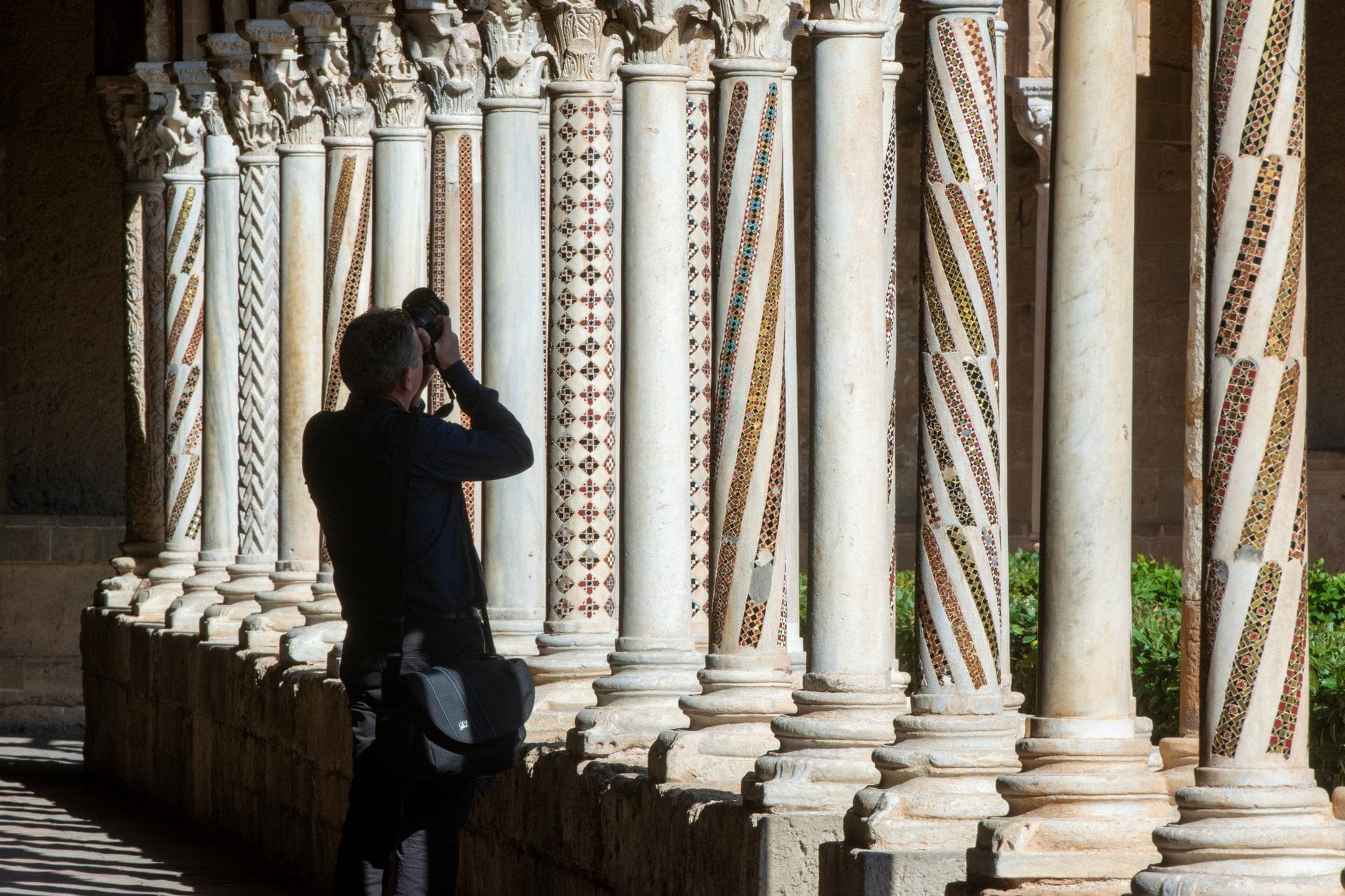 Besucher fotografiert die kunstvollen, mosaikverzierten Säulen des Kreuzgangs von Monreale im hellen Sonnenlicht, mit einem üppigen grünen Garten im Hintergrund.
