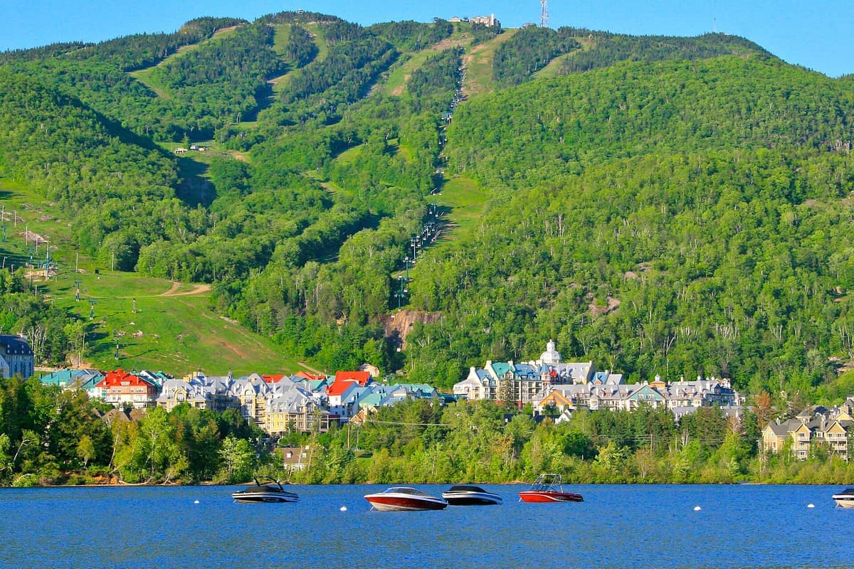 Boote treiben auf einem blauen See vor dem Dorf Mont-Tremblant, mit bewaldeten Bergen und Skipisten im Hintergrund.