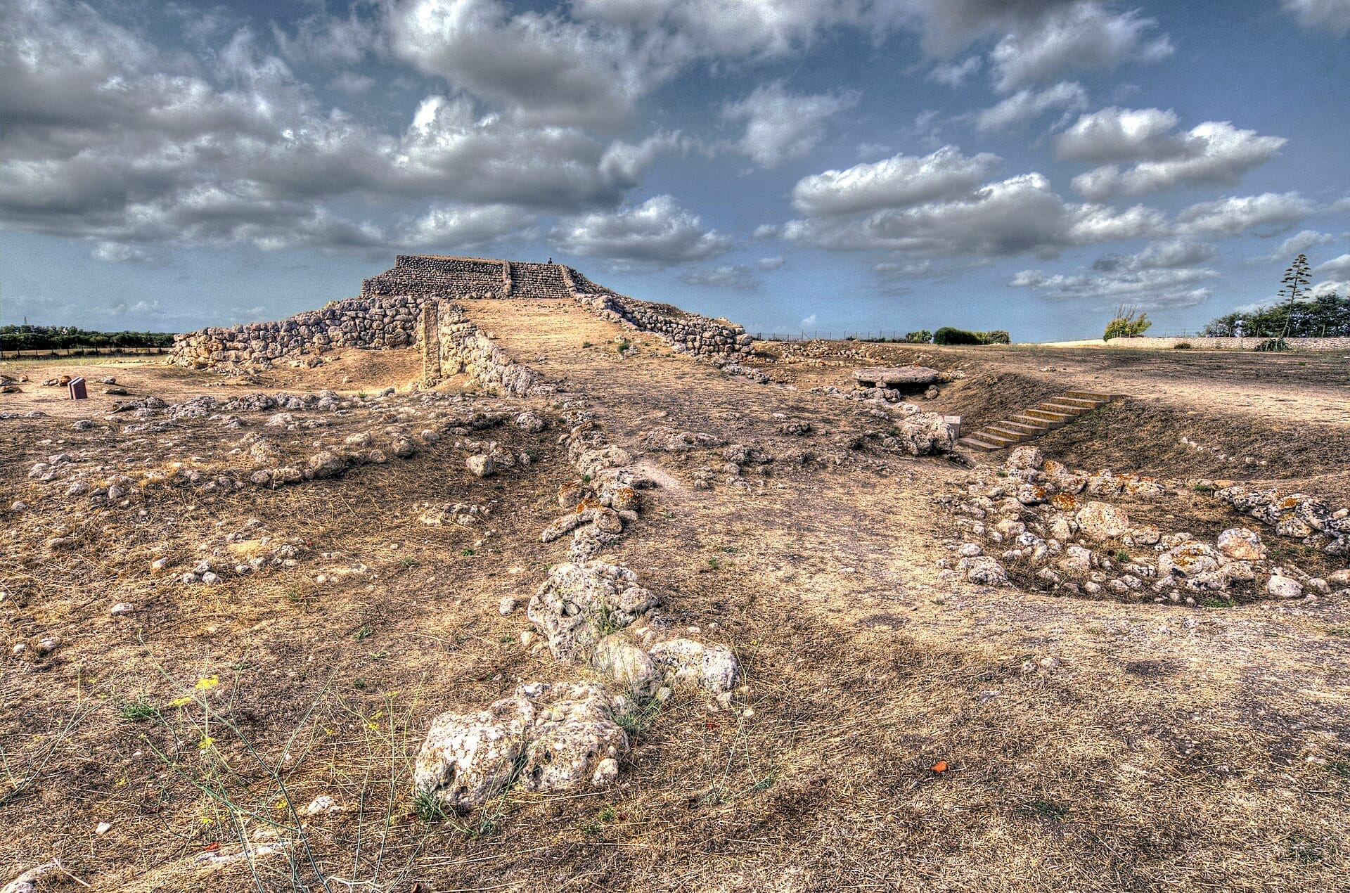 Steinrampe und Altarstruktur von Monte d'Accoddi unter einem dramatischen Himmel, umgeben von trockenem Gras und verstreuten Felsen auf Sardinien.