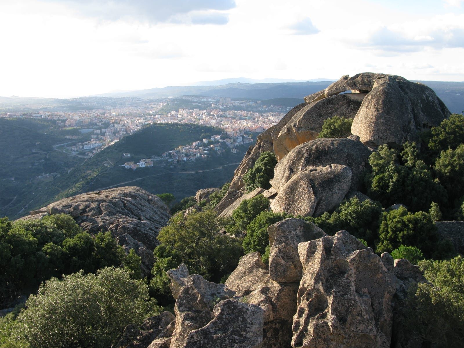 Aussicht vom Monte Ortobene mit schroffen Granitfelsen, grünem Wald und der Stadt Nuoro, die sich im fernen Tal unten erstreckt.