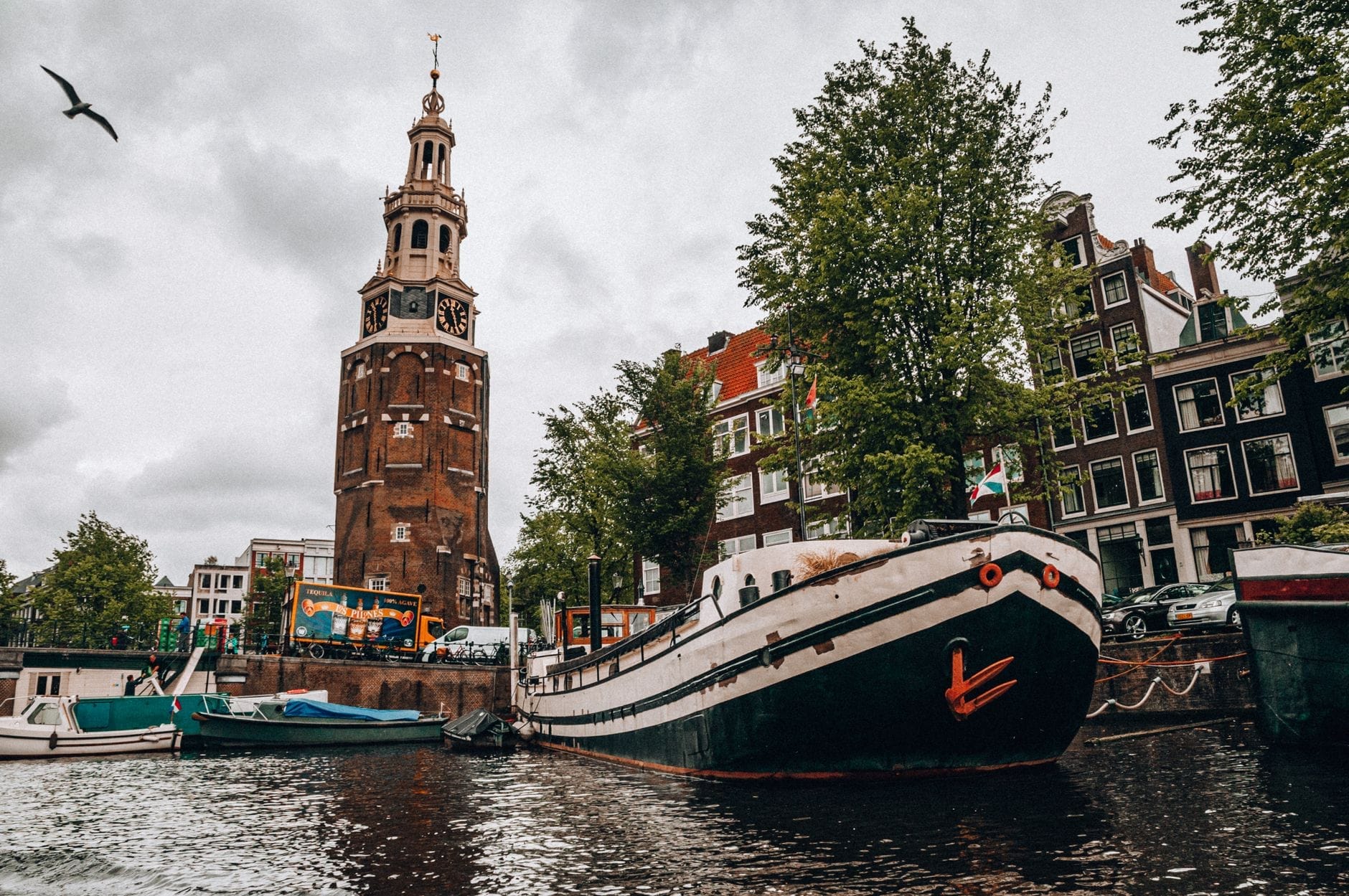 Vista de la Montelbaanstoren sobre el canal Oudeschans en Ámsterdam, con casas flotantes, árboles, fachadas junto al canal y un pájaro volando bajo un cielo nublado.