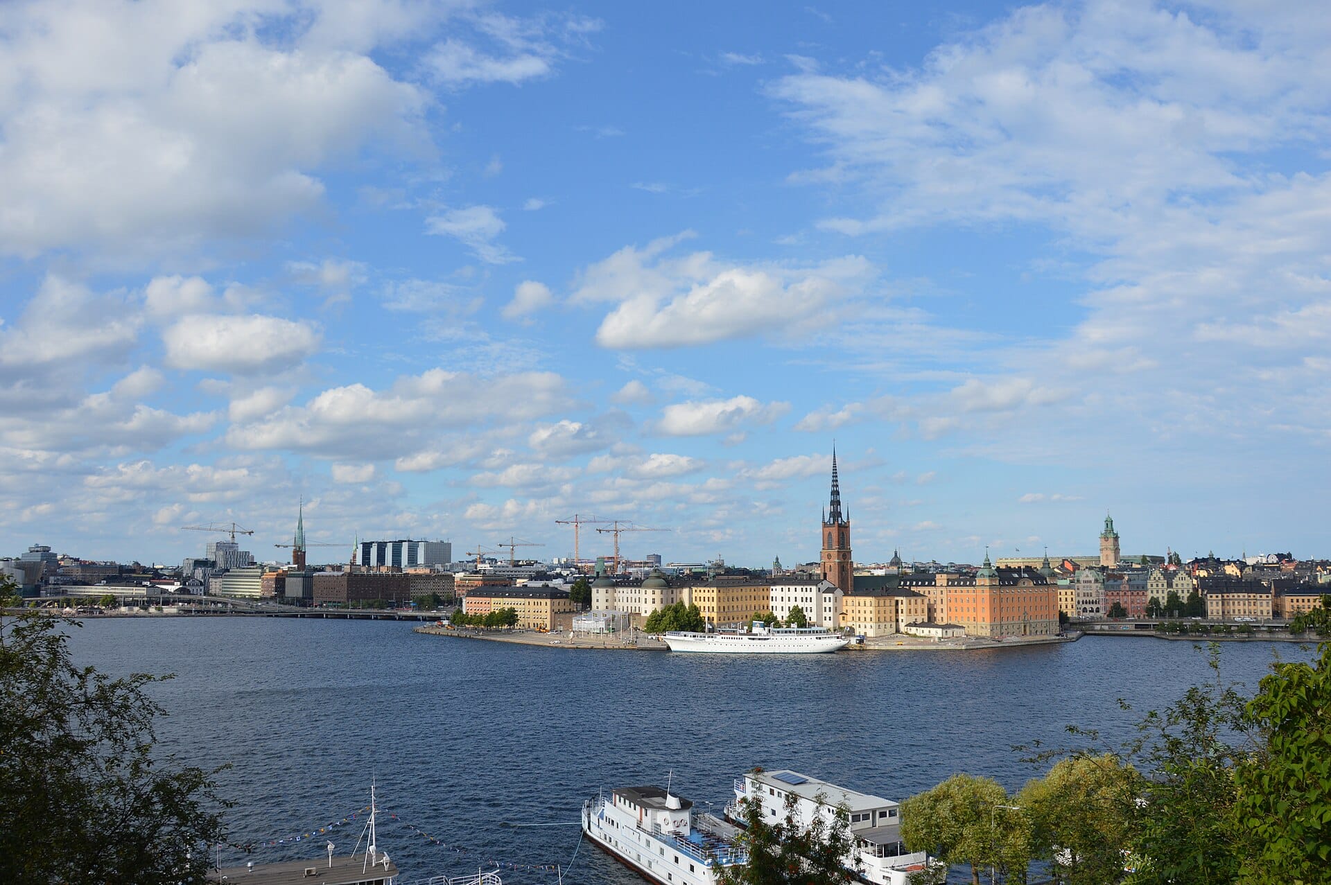 วิวพาโนรามาจาก Monteliusvägen มองเห็นทะเลสาบเมลเรน เมืองเก่า Gamla Stan โบสถ์ Riddarholmen และเมืองสตอกโฮล์มใต้ท้องฟ้าสีน้ำเงินมีเมฆกระจาย
