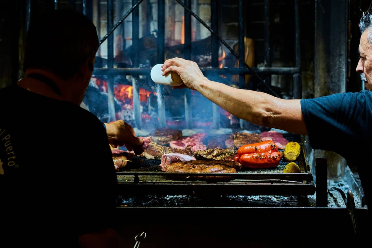 พ่อครัวสองคนกำลังเตรียม asado บนเตาย่างแบบเปิดที่มีเปลวไฟพร้อมเนื้อสัตว์และผักหลากหลายชนิด น่าจะอยู่ภายใน Mercado del Puerto ของ Montevideo