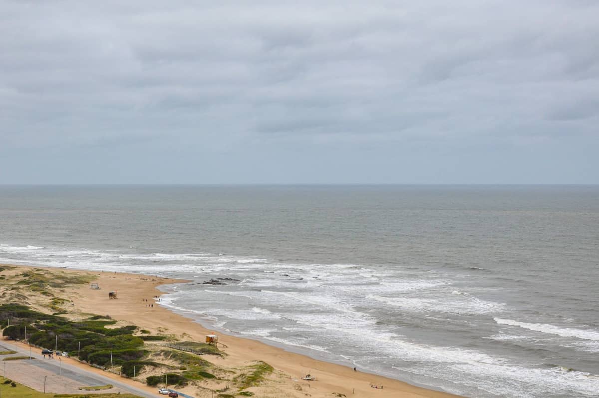 ชายหาดทรายกว้างพร้อมคลื่นมหาสมุทรและการพัฒนาบริเวณโดยรอบน้อย บ่งบอกถึงจุดหมายปลายทางนอกเมือง Montevideo ภายใต้ท้องฟ้ามีเมฆ