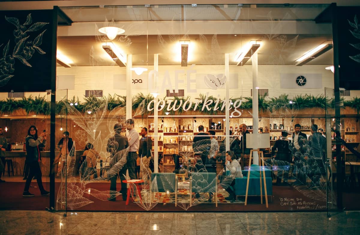 มุมมองผ่านกระจกด้านหน้าของพื้นที่ coworking และคาเฟ่ที่คึกคัก พร้อมผู้คนที่กำลังทำงานและพูดคุยสังสรรค์ภายใน