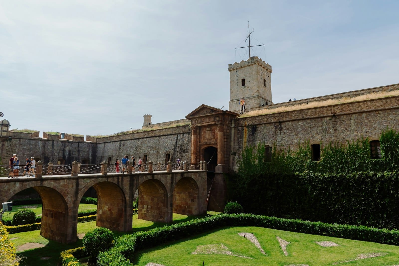 Visitors cross the stone arched bridge toward the main entrance of Montjuïc Castle, surrounded by manicured gardens and ancient fortress walls under a clear sky.