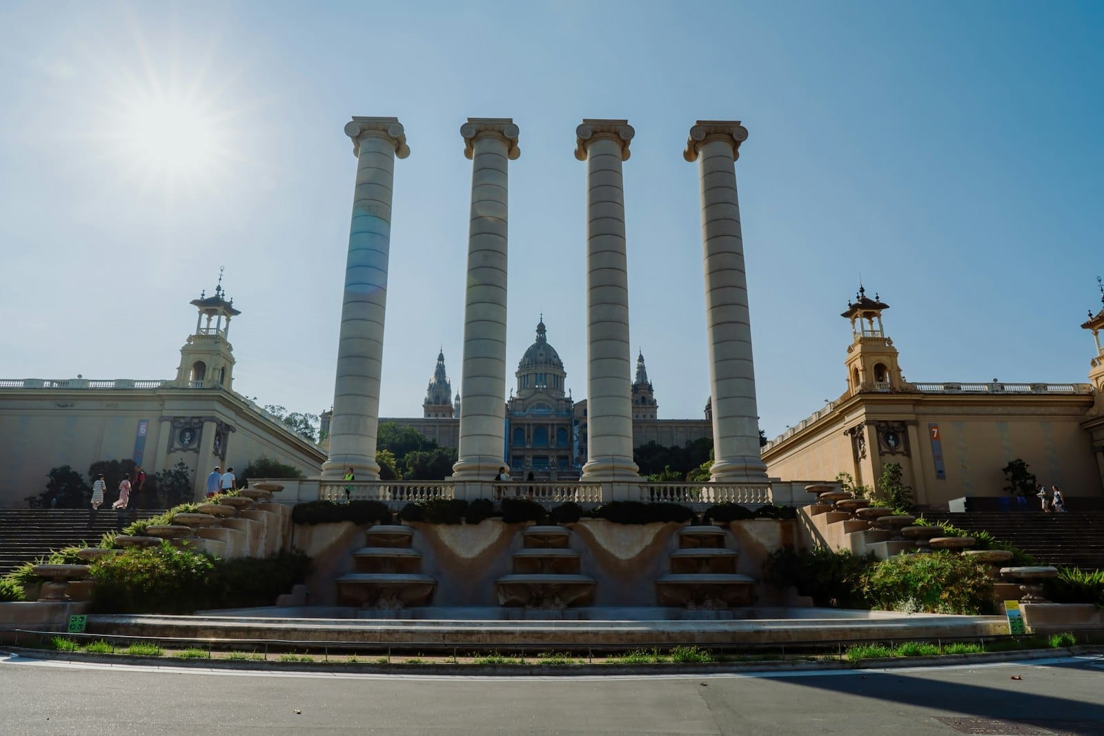 View of the four columns with the Magic Fountain and National Art Museum of Catalonia in the background, under a bright blue sky in Montjuïc, Barcelona.