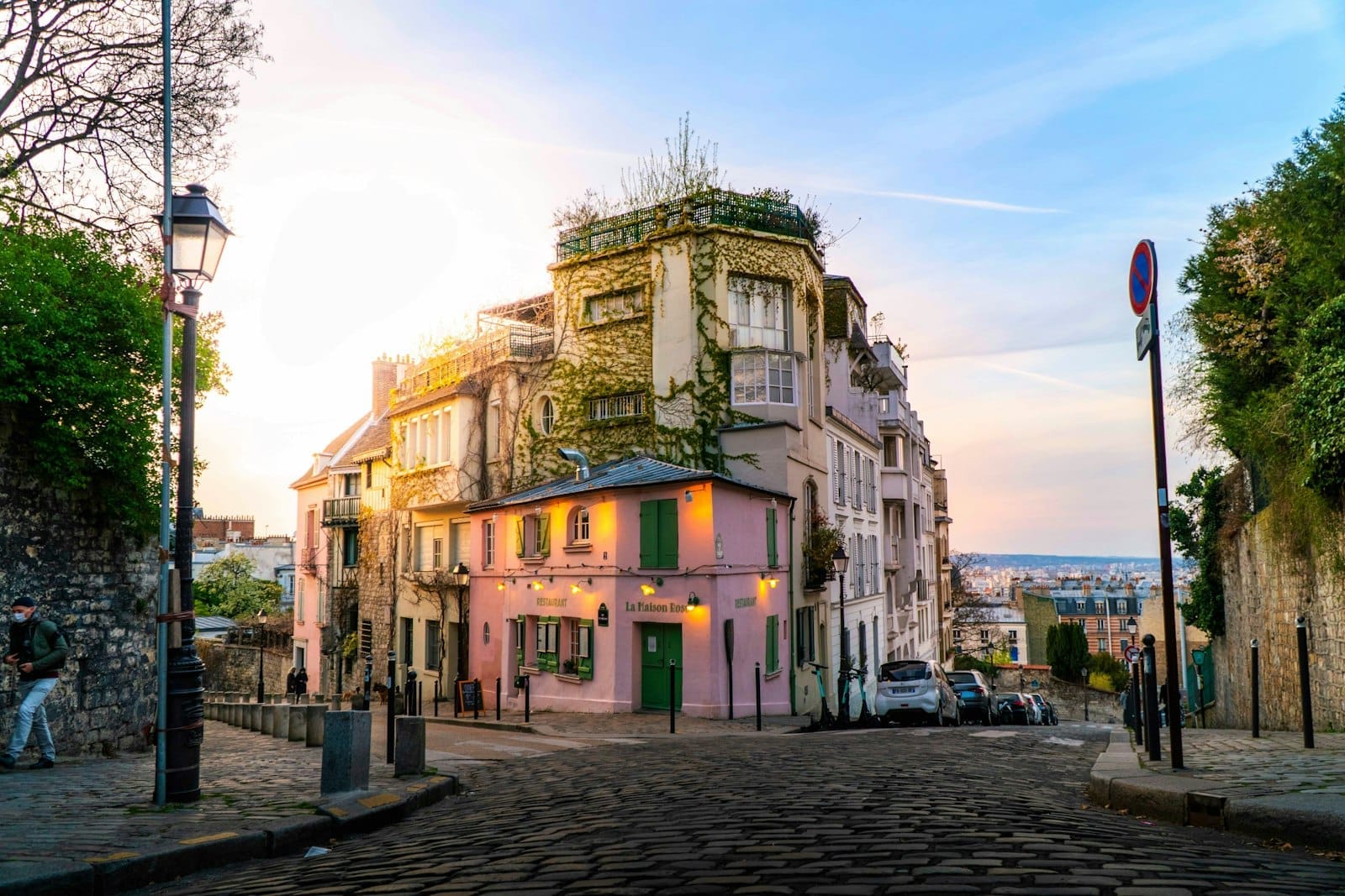 Un incantevole vicolo acciottolato di Montmartre con edifici ricoperti di viti, facciate pastello e un caffè rosa nella luce soffusa della sera, che cattura l'atmosfera bohémien parigina.