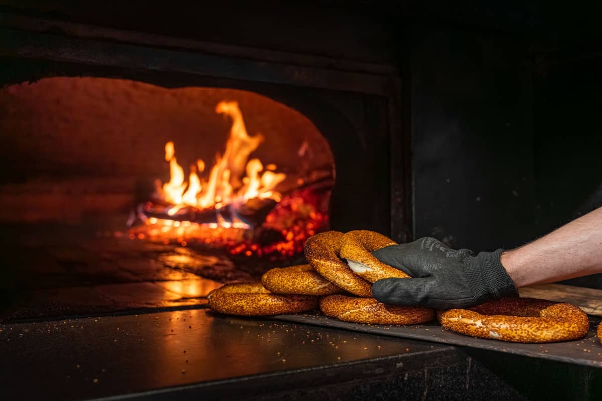Bagels frescos de Montreal sobre uma pá entrando em um forno a lenha com chamas visíveis ao fundo.