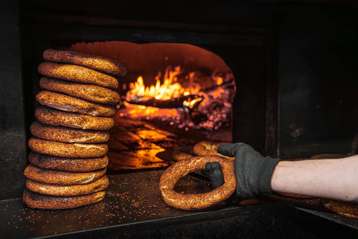 Uma pilha de bagels de gergelim de Montreal frescos ao lado de um forno a lenha, com uma mão enluvada segurando um bagel quente, mostrando o processo de panificação tradicional.