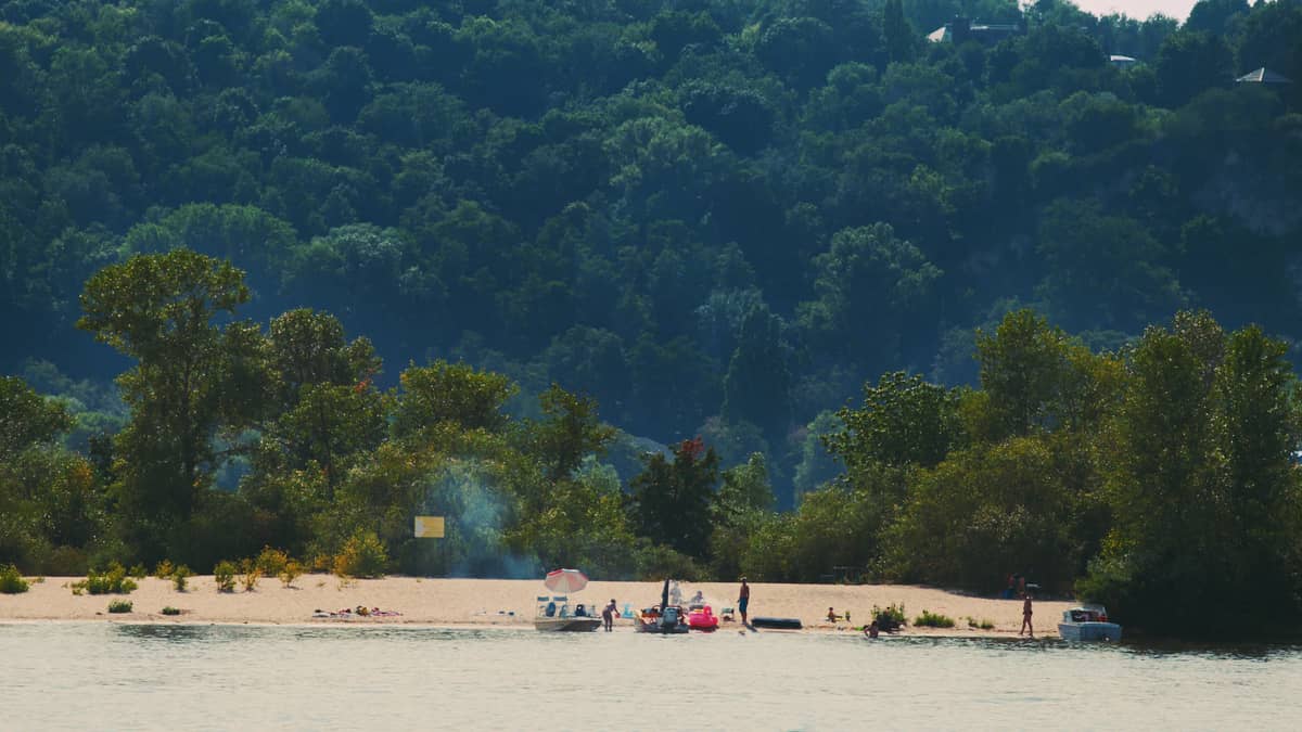 Small sandy beach with people and boats on a wooded river island, seen from across the water on a sunny summer day