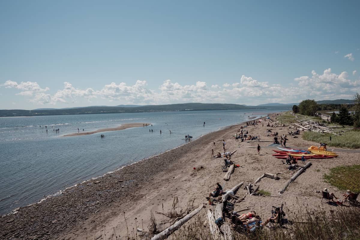 Ein breiter Sandstrand an einem Süßwassersee bei Montréal mit vereinzelten Sonnenbadenden, Kajaks und Treibholz, umgeben von Wasser und grünen Hügeln in der Ferne.