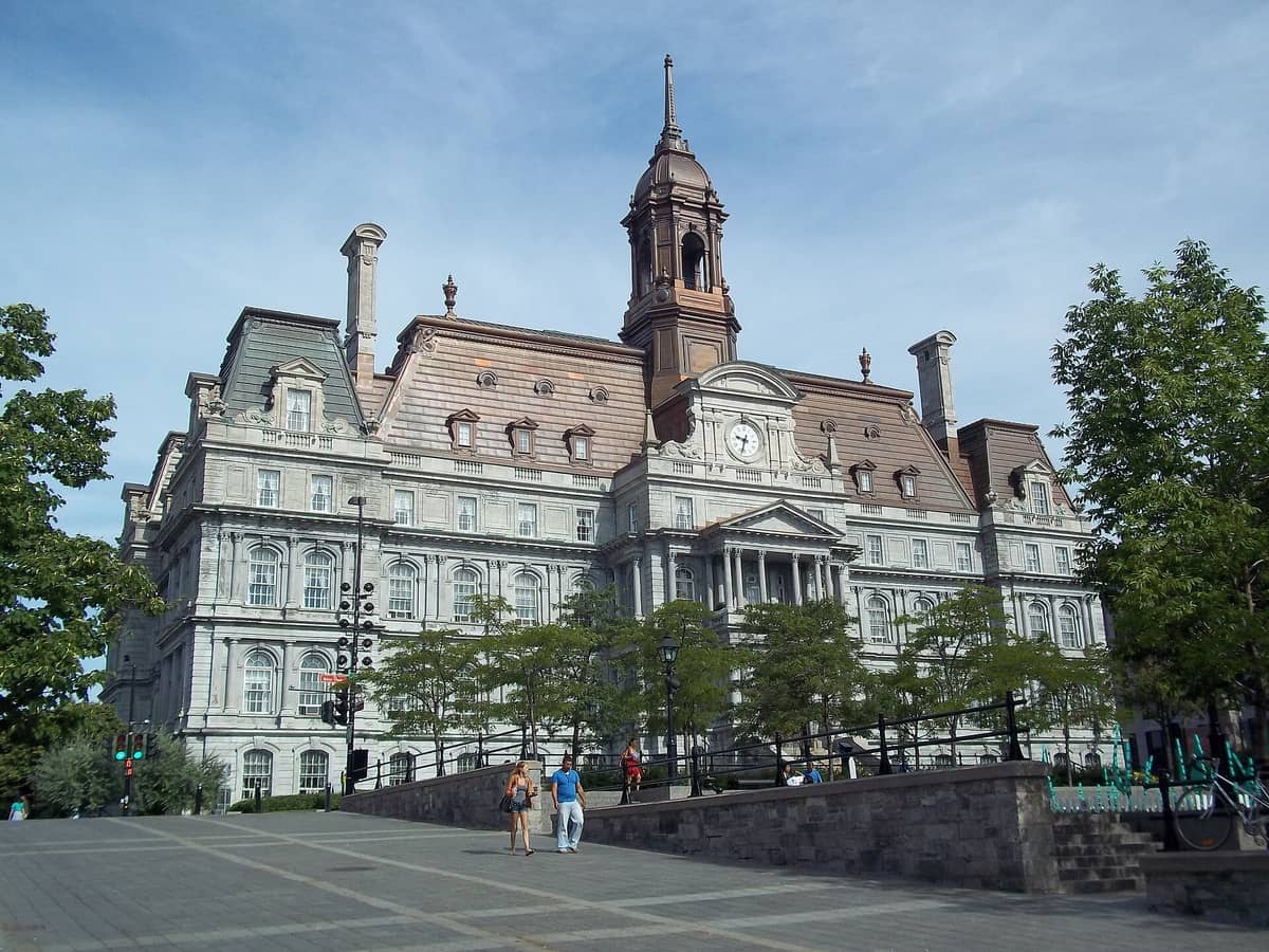 Das Montrealer Rathaus mit seiner verzierten Fassade und dem hohen Uhrturm, von einem gepflasterten Platz mit Bäumen und mehreren vorbeigehenden Fußgängern aus gesehen.