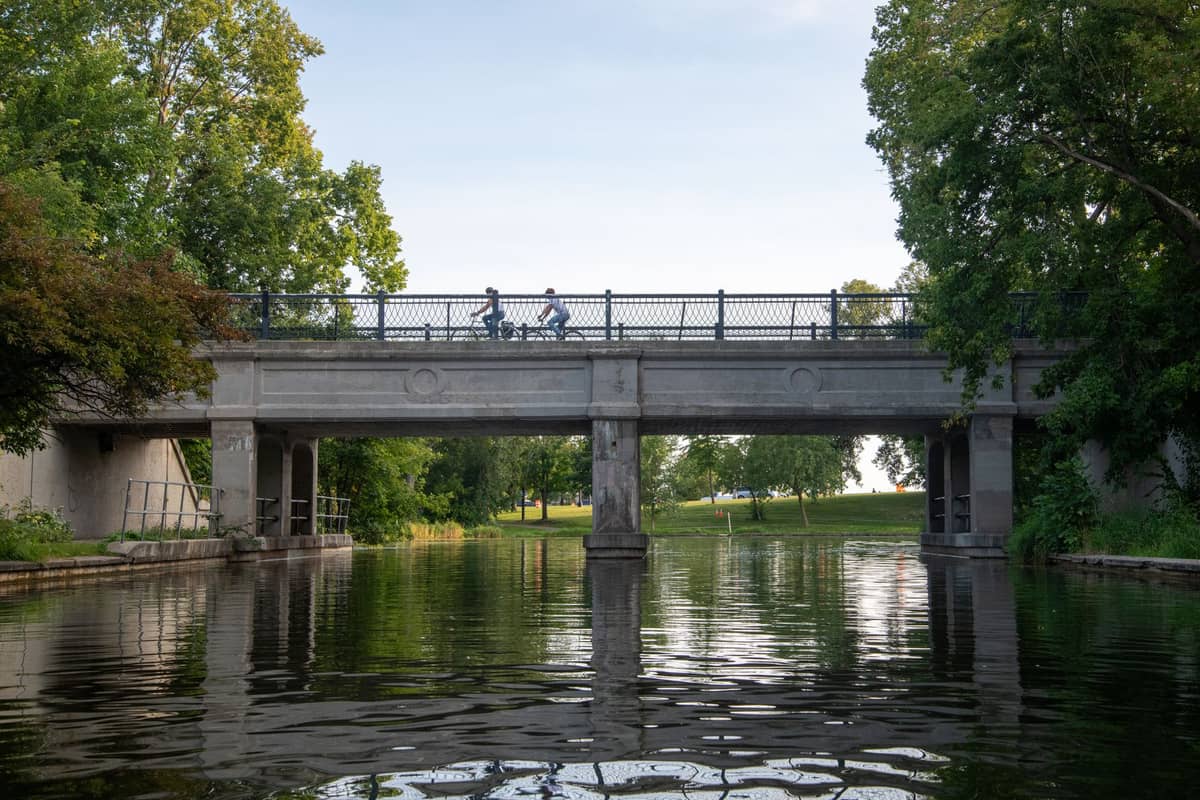 นักปั่นจักรยานสองคนปั่นข้ามสะพานเหนือคลอง Lachine ในเมือง Montreal รายล้อมด้วยต้นไม้เขียวขจีและน้ำที่สงบด้านล่าง