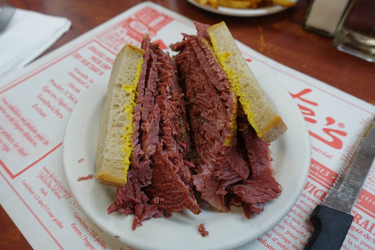 Ein klassisches Montreal Smoked Meat Sandwich auf Roggenbrot mit Senf, serviert auf einem weißen Teller an einem Delicatessen-Tisch.