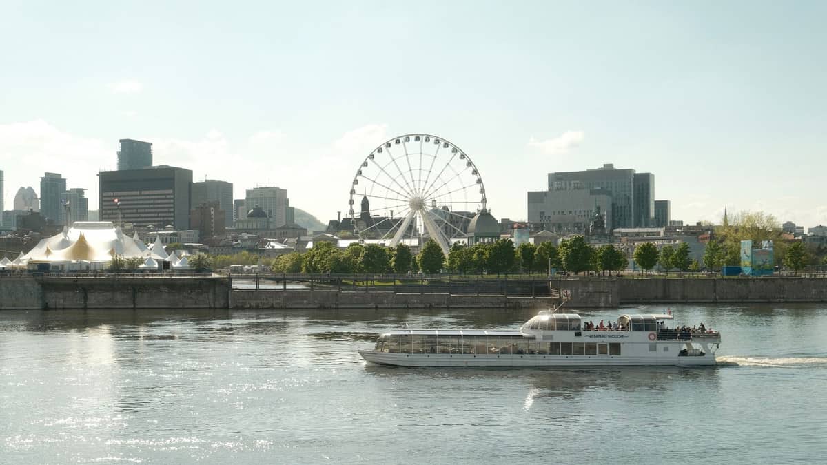 Blick auf den Alten Hafen von Montréal mit Skyline, dem Riesenrad La Grande Roue und einem Ausflugsboot auf dem Fluss an einem sonnigen Tag.
