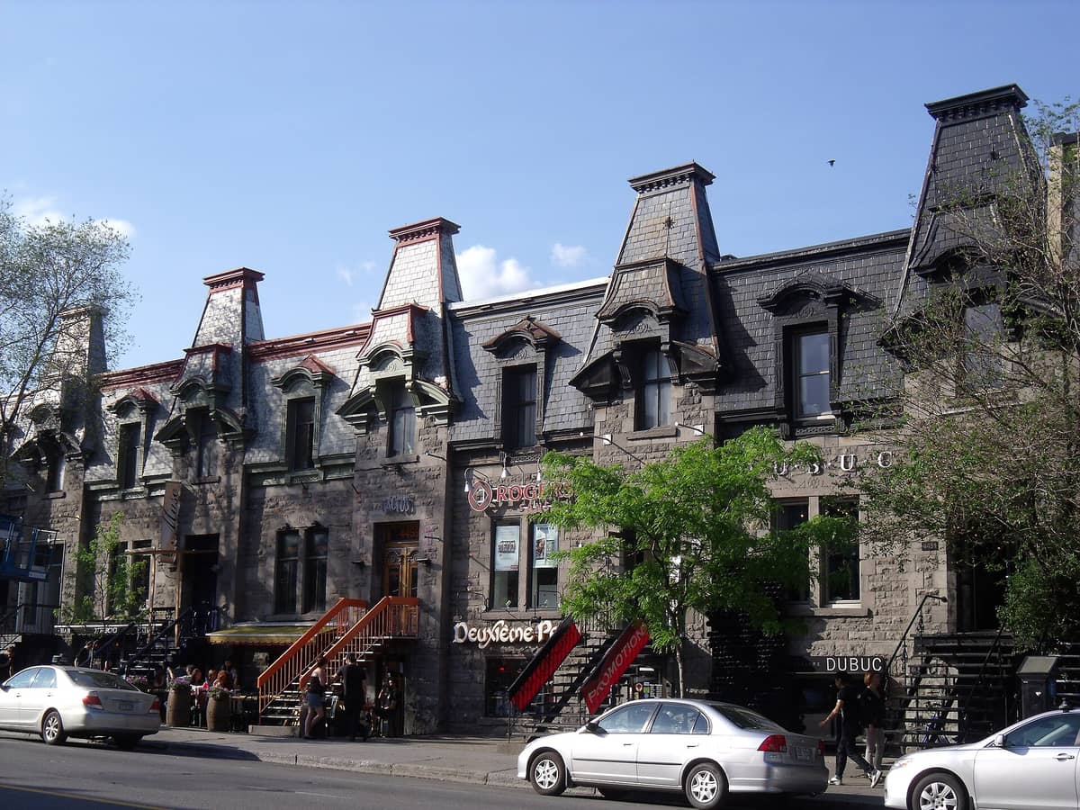 บ้านแถวสไตล์เอกลักษณ์ของ Montreal ที่มีหลังคา mansard ชันและหน้าต่าง dormer ประดับประดาอย่างสวยงาม บนถนนในเมืองที่คึกคักพร้อมรถที่จอดอยู่และผู้คนนั่งโต๊ะกลางแจ้ง