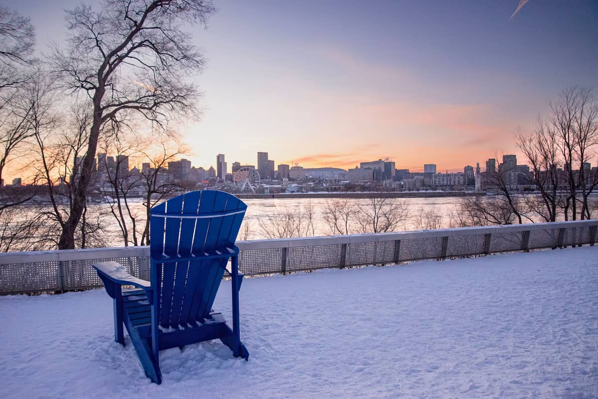 Schneebedecktes Flussufer mit einem großen blauen Stuhl, der bei Sonnenuntergang auf die Skyline von Montreal blickt, kahle Bäume im Vordergrund.