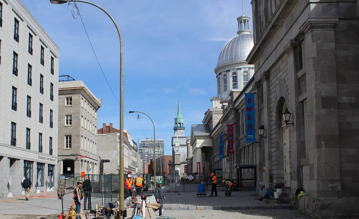 Straße im Alten Montreal mit der historischen Kuppel des Marché Bonsecours und dem Kirchturm, Steingebäuden und Menschen, die in der Gegend spazieren.