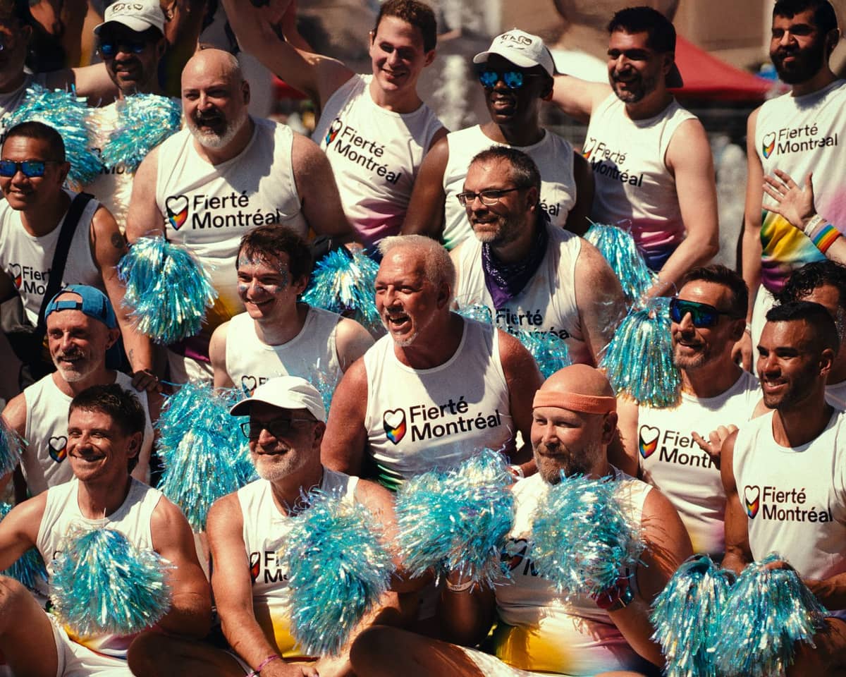 Eine Gruppe von Menschen in Fierté-Montréal-Shirts mit Pompons bei der Montréal Pride – draußen, bei strahlendem Sonnenschein.