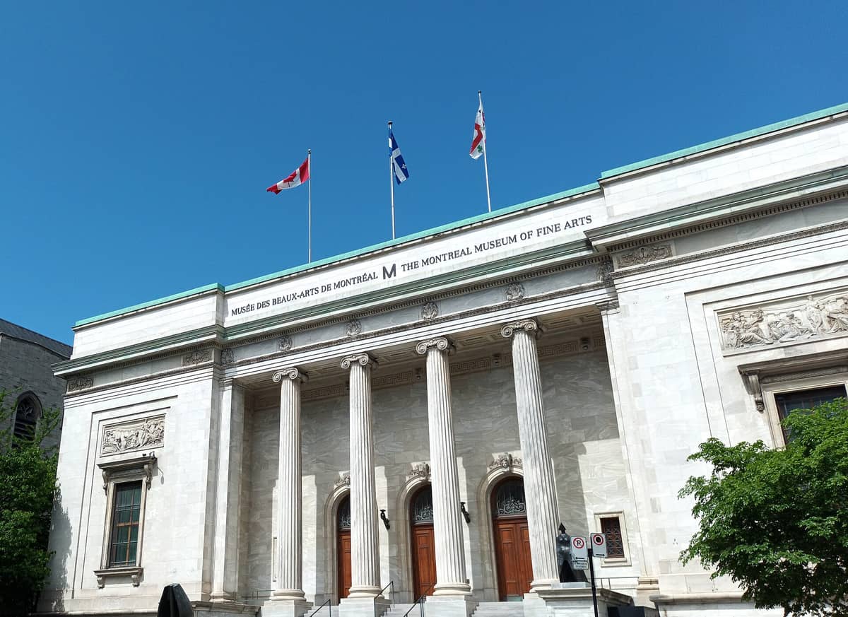 Der Haupteingang des Montrealer Museums der Schönen Künste mit dem Namen auf Englisch und Französisch, vier Säulen und drei Flaggen darüber, an einem sonnigen Tag.