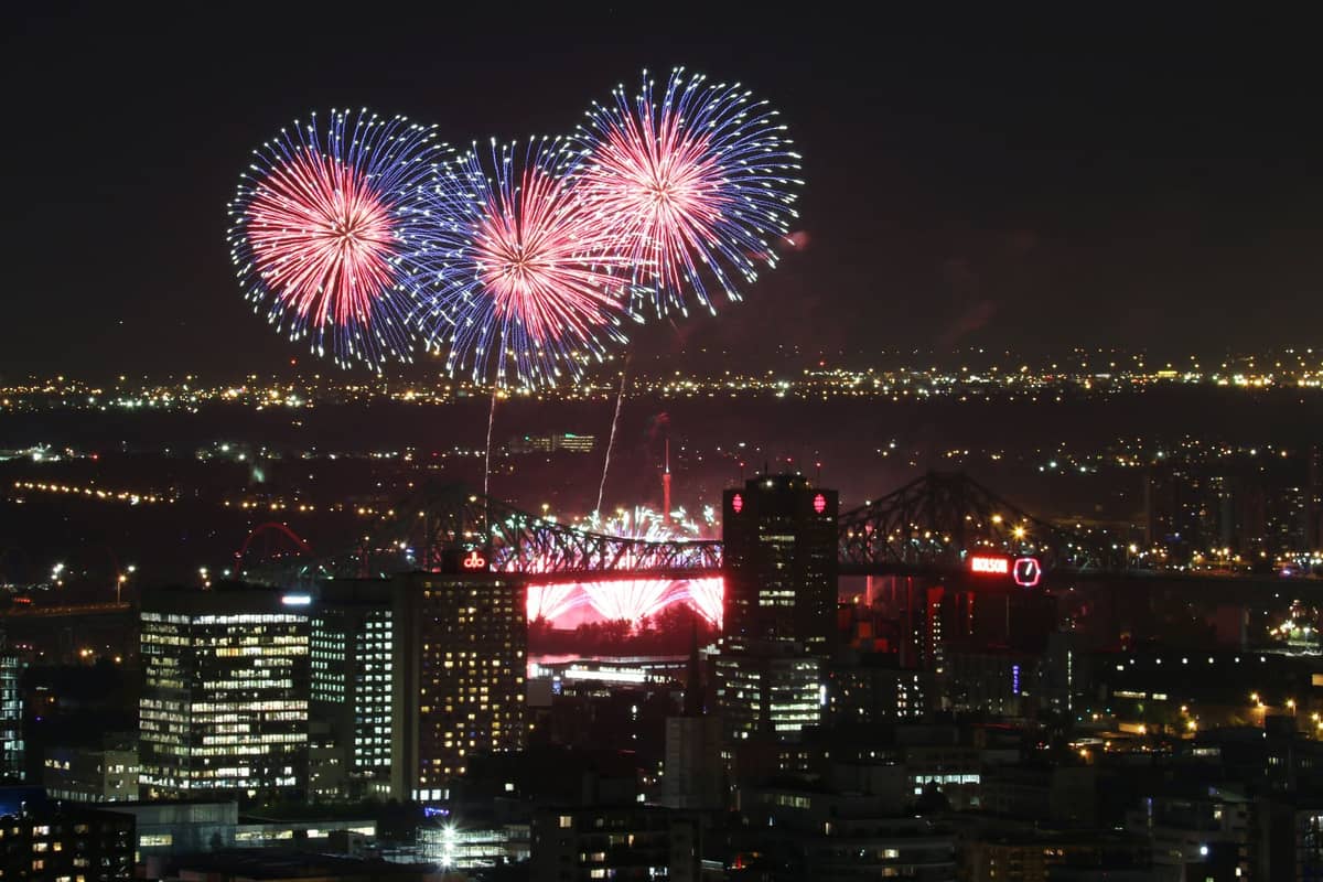 Vista notturna del centro di Montreal con fuochi d'artificio che esplodono sopra lo skyline della città e il illuminato Ponte Jacques-Cartier sullo sfondo.