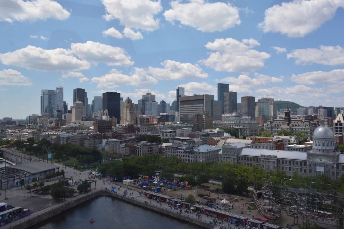 Panoramablick auf die Skyline Montréals tagsüber, mit dem Alten Montréal und dem Hafenbereich unter einem blauen Himmel mit weißen Wolken.