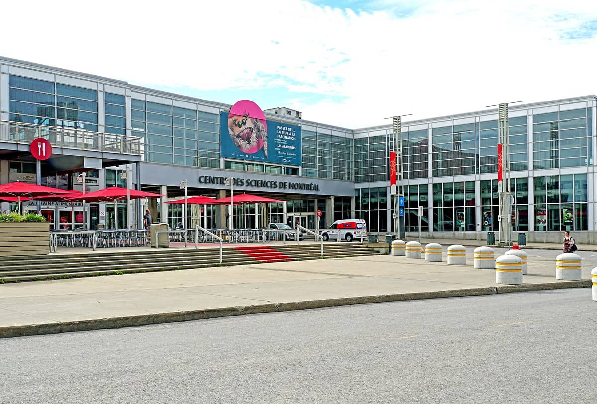 Außenansicht des Montreal Science Centre mit Glasfassade, roten Sonnenschirmen und dem Eingangsschild am King-Edward-Kai im Alten Hafen von Montréal.