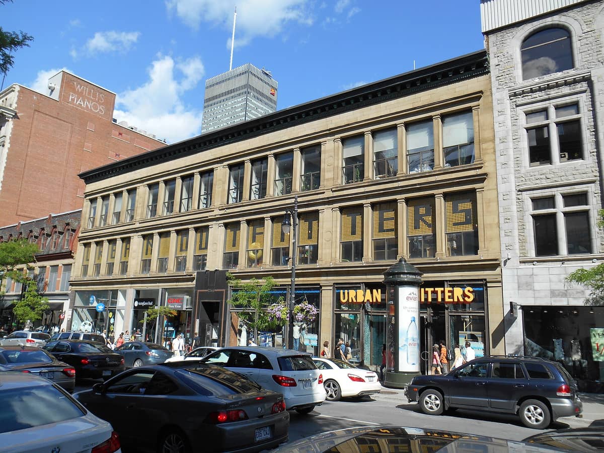 ถนน Sainte-Catherine ที่คึกคักในมอนทรีออลเต็มไปด้วยรถยนต์และผู้คน โดยมองเห็นหน้าร้านประวัติศาสตร์อย่าง Urban Outfitters และอาคาร Willis Pianos